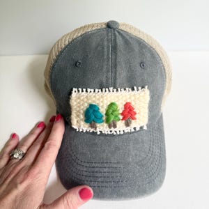 May include: Gray trucker hat with a beige mesh back and a decorative patch featuring three colorful trees. The trees are teal, green, and red, stitched onto a cream-colored background.