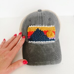 May include: A gray baseball cap with a woven patch featuring a mountain landscape design in shades of blue, orange, yellow, and white. The cap has a curved brim and a tan mesh back.