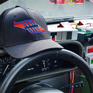 Może przedstawiać: Czarna czapka typu trucker z czerwono-niebieską grafiką leży na desce rozdzielczej samochodu. Projekt czapki przedstawia stylizowaną strzałkę. Wnętrze samochodu obejmuje wskaźniki, kierownicę i wyświetlacz podróży w czasie.