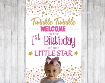 May include: A vertical sign with the text "Twinkle Twinkle Welcome to the 1st Birthday of our Little Star" in gold and pink. The sign has a background of gold, pink, and silver stars. A baby with a pink bow is at the bottom of the sign.