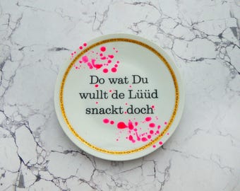Può includere: Un piatto rotondo in ceramica bianca con un bordo decorativo dorato e il testo nero Do wat Du wullt de Lüüd snackt doch. Il piatto è decorato con schizzi di vernice rosa brillante sulla superficie, creando un netto contrasto visivo.