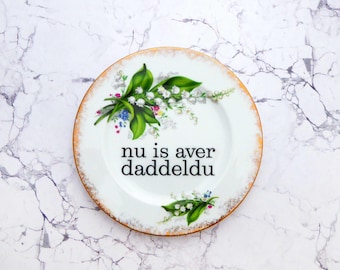 Può includere: Un piatto in ceramica bianca con bordo dorato, caratterizzato da un motivo floreale con foglie verdi e piccoli fiori bianchi di mughetto. Al centro del piatto è presente il testo stampato in nero "nu is aver daddeldu" in un semplice carattere serif.