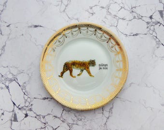 Puede incluir: Un plato de cerámica blanca con una ilustración central de un tigre caminando y el texto "nützt ja nix". El borde está decorado con un intrincado patrón de filigrana dorada que consiste en guirnaldas, puntos y motivos de hojas.