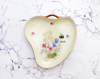 Puede incluir: Un plato de cerámica color crema con forma de corazón y un asa dorada metálica. El centro presenta un ramo de flores en rosa, azul y amarillo, un pequeño abejorro y la palabra "yeah" impresa en letras minúsculas negras.