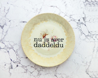 Puede incluir: Un plato redondo de cerámica con un centro de color crema que presenta una ilustración floral y el texto negro "nu is aver daddeldu". El borde está decorado con un acabado festoneado y finas líneas verticales doradas que irradian.