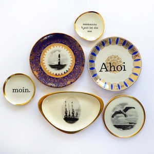 K&ouml;nnte beinhalten: F&uuml;nf dekorative Keramikteller mit goldenen R&auml;ndern und blau-wei&szlig;en Designs. Die Teller zeigen die Worte "Moin", "Ahoi" und "Seemanns gut ist die See".