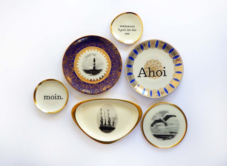 K&ouml;nnte beinhalten: F&uuml;nf dekorative Keramikteller mit goldenen R&auml;ndern und blau-wei&szlig;en Designs. Die Teller zeigen die Worte "Moin", "Ahoi" und "Seemanns gut ist die See".