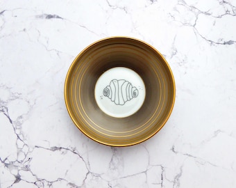 Può includere: Una ciotola rotonda in ceramica con interno color oro metallizzato caratterizzato da scanalature circolari concentriche. Il centro della ciotola presenta un semplice disegno a linee nere di un cornetto su sfondo bianco, circondato da piccoli punti neri.