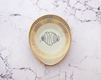 Può includere: Un poggia cucchiaio in ceramica color crema a forma di goccia, caratterizzato da un bordo dipinto in oro e da un motivo radiante di trattini dorati lungo il bordo interno. Un disegno a linee nere di un cornetto è centrato al centro.