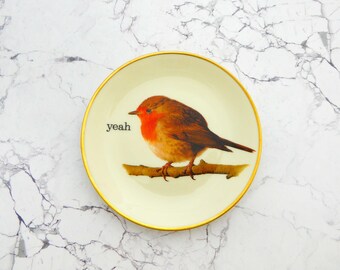Puede incluir: Un plato redondo de cerámica con base color crema y un borde dorado fino. El centro presenta una ilustración detallada de un petirrojo posado en una rama marrón. A la izquierda del ave, la palabra "yeah" está impresa en una fuente negra.