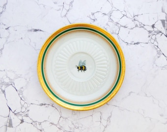 Puede incluir: Un platillo redondo de porcelana con una ilustración central de un abejorro. El platillo tiene un centro blanco con estrías radiales en relieve, rodeado por una banda verde oscuro y un borde dorado ancho y texturizado con puntos.