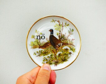 Puede incluir: Un pequeño plato redondo de porcelana blanca con un borde dorado. El centro presenta una ilustración colorida de un faisán en un campo de hierba con árboles y flores blancas. El texto en minúsculas "nö" está impreso en negro a la izquierda.