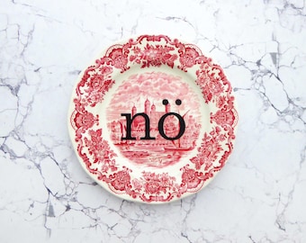 Puede incluir: Un plato redondo de cerámica con un patrón de transferencia en rojo y blanco que muestra un castillo y bordes florales. La palabra "nö" está impresa en letras minúsculas negras grandes en el centro del plato, sobre la escena detallada.