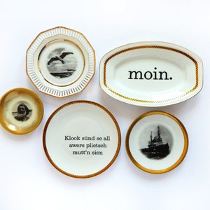 K&ouml;nnte beinhalten: F&uuml;nf wei&szlig;e Keramikteller mit goldenen R&auml;ndern und schwarzen Designs. Ein Teller hat eine M&ouml;we, einer ein Schiff, einer ein Portr&auml;t, einer das Wort "moin." und einer den Text "Klook s&uuml;ind se all awers plietsch mutt'n sien."