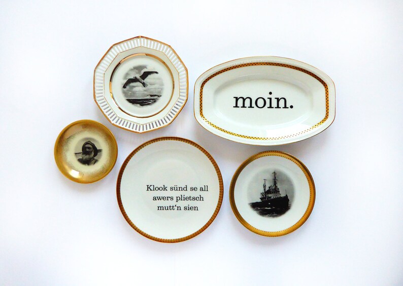 K&ouml;nnte beinhalten: F&uuml;nf wei&szlig;e Keramikteller mit goldenen R&auml;ndern und schwarzen Designs. Ein Teller hat eine M&ouml;we, einer ein Schiff, einer ein Portr&auml;t, einer das Wort "moin." und einer den Text "Klook s&uuml;ind se all awers plietsch mutt'n sien."