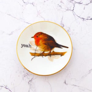 Puede incluir: Un plato pequeño redondo de cerámica blanca con un borde dorado. El centro presenta una ilustración detallada de un petirrojo posado en una rama marrón con hojas claras. La palabra "yeah" está impresa en letras minúsculas negras a la izquierda.