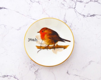 Puede incluir: Un plato pequeño redondo de cerámica blanca con un borde dorado. El centro presenta una ilustración detallada de un petirrojo posado en una rama marrón con hojas claras. La palabra "yeah" está impresa en letras minúsculas negras a la izquierda.