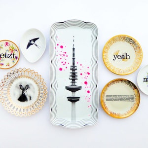K&ouml;nnte beinhalten: Ein Set aus sieben dekorativen Tellern mit verschiedenen Designs. Die Teller zeigen eine Schwarzwei&szlig;illustration eines Turms, eine Schwarzwei&szlig;illustration eines Kaninchens, eine Schwarzwei&szlig;illustration eines Vogels und die W&ouml;rter "jetzt", "yeah", "moin".