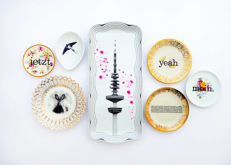 K&ouml;nnte beinhalten: Ein Set aus sieben dekorativen Tellern mit verschiedenen Designs. Die Teller zeigen eine Schwarzwei&szlig;illustration eines Turms, eine Schwarzwei&szlig;illustration eines Kaninchens, eine Schwarzwei&szlig;illustration eines Vogels und die W&ouml;rter "jetzt", "yeah", "moin".