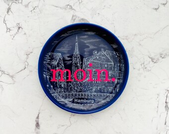 Può includere: Un piatto rotondo in ceramica blu scuro con un'illustrazione a linee bianche della chiesa di San Nicola ad Amburgo, in Germania. La parola "moin." è stampata in rosa brillante al centro, con "Hamburg" e "NICOLAIKIRCHE" scritti sotto.