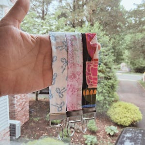 May include: Three fabric keychains with different patterns are displayed. One is white with blue bows, another is pink with floral designs, and the third is a mix of black, pink, and orange. Each keychain has a silver-colored metal clasp.