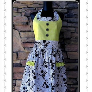 Puede incluir: Delantal para adultos 'Lime Rickey' con corpiño amarillo y falda estampada con círculos blancos y negros. Tiene tres botones negros y detalles verde lima en los bolsillos. El delantal se exhibe en un maniquí.