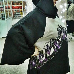 May include: A black hooded poncho with a beige front panel featuring a graphic print of purple and black feathers arranged in a curved pattern. The garment has wide sleeves and a solid black back and hood, providing a loose, draped fit.