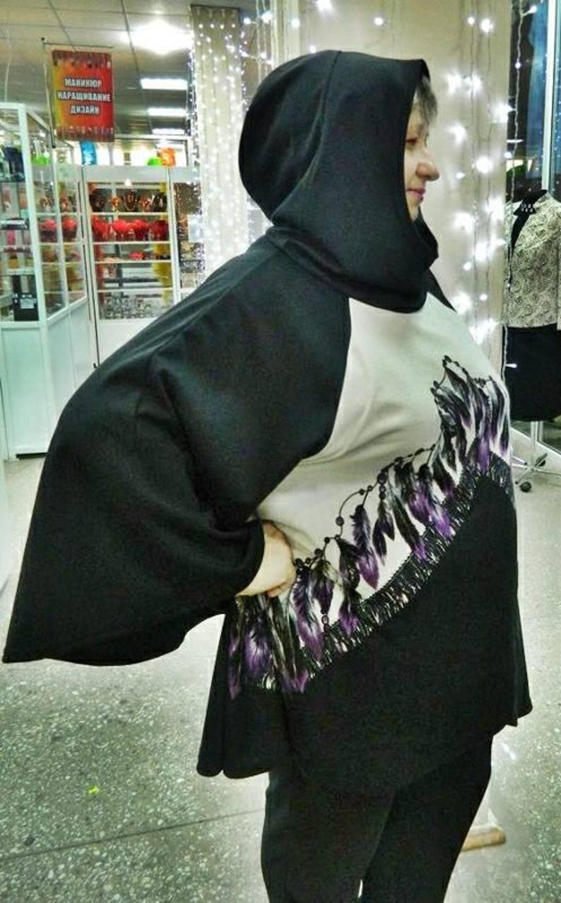 May include: A black hooded poncho with a beige front panel featuring a graphic print of purple and black feathers arranged in a curved pattern. The garment has wide sleeves and a solid black back and hood, providing a loose, draped fit.