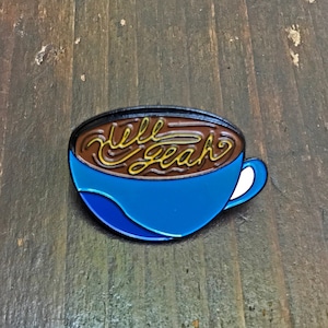 May include: Blue enamel pin shaped like a coffee cup with brown coffee and the words "hell yeah" written in yellow in the coffee.