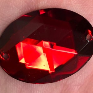May include: A faceted, oval-shaped, deep red crystal with two small holes for stringing. The crystal's surface reflects light, creating a brilliant display of color and geometric patterns. This decorative element is suitable for jewelry making or craft projects.