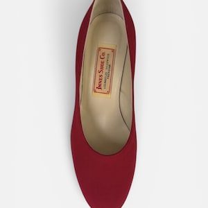 May include: A deep red pump shoe with a rounded toe and a smooth exterior finish. The interior lining is a light beige color and features a rectangular label with the text Innes Shoe Co. Los Angeles Hollywood Pasadena printed in red ink.
