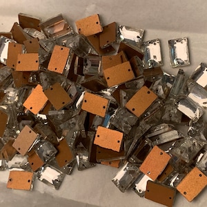 May include: A collection of clear, rectangular rhinestones with a faceted cut, interspersed with small, brown, rectangular pieces. Each brown piece has two small holes. The rhinestones are scattered across a light-colored surface.