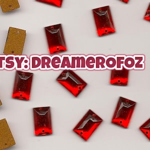 May include: Scattered across a light gray surface are several rectangular, ruby-red glass rhinestones and a few gold-colored tags. The Etsy shop name, DREAMEROFOZ, is displayed in a pink and white banner.