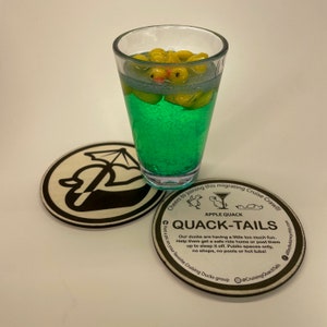 May include: A clear glass filled with a green liquid and yellow rubber duckies. Two round wooden coasters are in the foreground. One coaster has a black and white logo, and the other has text that reads "Apple Quack Quack-Tails".
