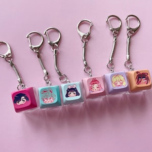 May include: Six keycap keychains featuring colorful square plastic caps with printed anime-style character faces. Each keychain includes a silver metal ring and a flexible coiled spring connector attached to the top of the individual keycap.
