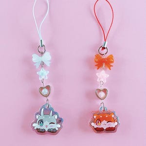 以下が含まれることがあります： ピンク色の背景に、2つのチャームキーホルダー。1つは白い紐、白いリボン、星、ハート、漫画の恐竜。もう1つは赤い紐、オレンジ色のリボン、星、ハート、漫画の猫です。
