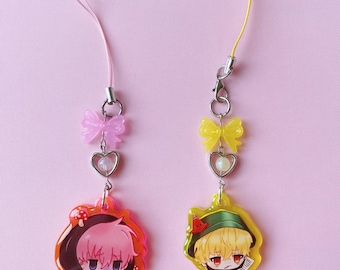 May include: Two phone charms featuring anime-style character heads. The left charm has a pink-haired character, a pink bow, and a heart-shaped bead. The right charm has a blonde character in a green hat, a yellow bow, and a heart-shaped bead.