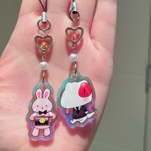 May include: Two iridescent acrylic charms with black cords. One charm features a pink bunny holding a television with a smiley face. The other charm depicts a cartoon character in a suit. Both charms have heart-shaped accents and pearl beads.
