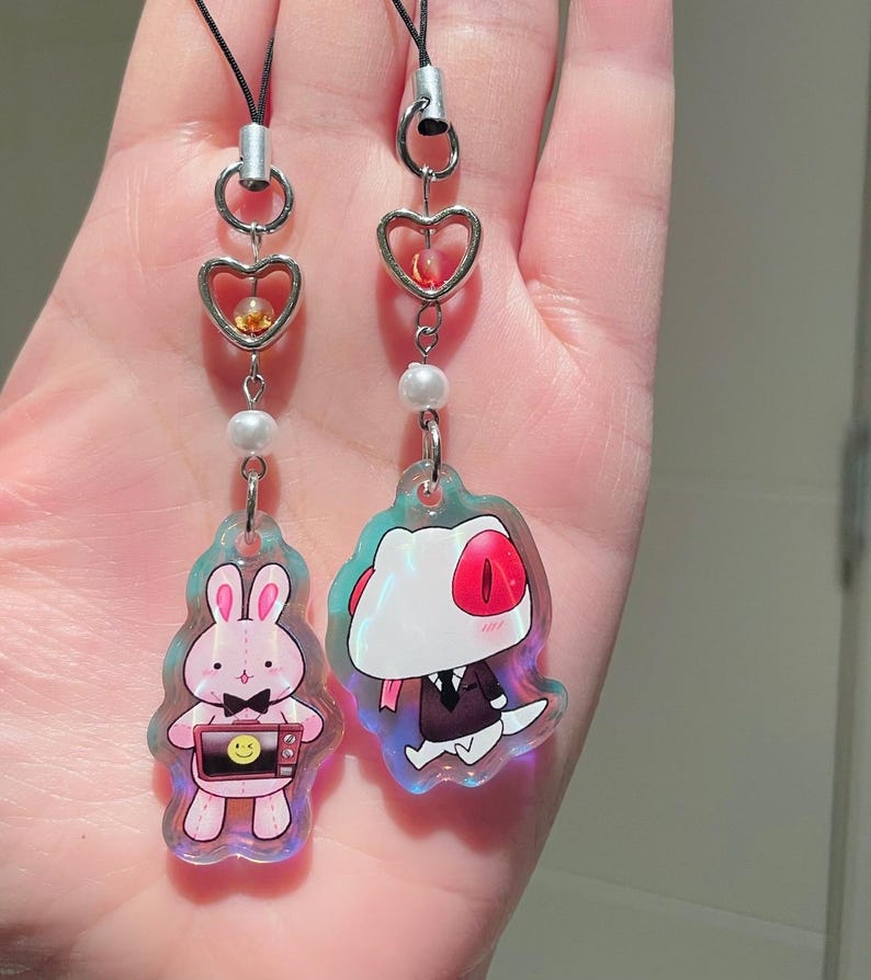 May include: Two iridescent acrylic charms with black cords. One charm features a pink bunny holding a television with a smiley face. The other charm depicts a cartoon character in a suit. Both charms have heart-shaped accents and pearl beads.