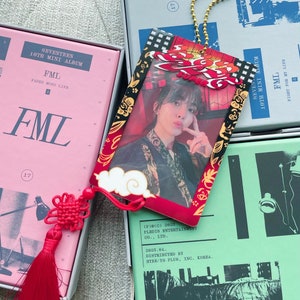 May include: A red and black keychain with a photo of a person in a red and black patterned shirt. The keychain has a gold chain and a red tassel. The text "FML" is printed on the keychain. The text "(P)(C) 2025 PLEDIS ENTERTAINMENT CO., LTD. 2025.04. DISTRIBUTED BY HYBE/YG PLUS, INC. KOREA." is printed on the back of the keychain.
