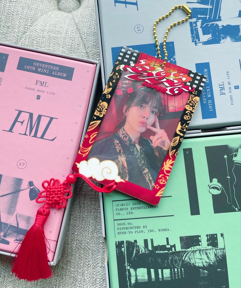 May include: A red and black keychain with a photo of a person in a red and black patterned shirt. The keychain has a gold chain and a red tassel. The text "FML" is printed on the keychain. The text "(P)(C) 2025 PLEDIS ENTERTAINMENT CO., LTD. 2025.04. DISTRIBUTED BY HYBE/YG PLUS, INC. KOREA." is printed on the back of the keychain.