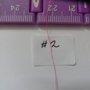 May include: A close-up of a pink thread with a white label that says "#2" on it. The thread is hanging from a purple ruler.