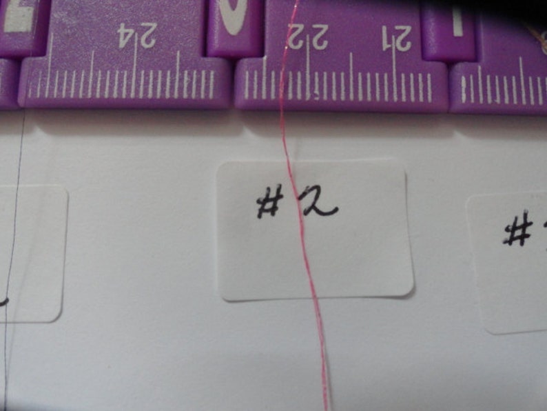 May include: A close-up of a pink thread with a white label that says "#2" on it. The thread is hanging from a purple ruler.