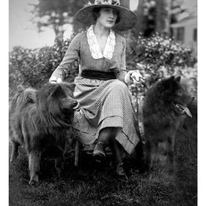 Könnte beinhalten: Eine Frau in einem Strohhut und einem karierten Kleid sitzt mit zwei schwarzen Hunden. Die Frau hält eine Leine in ihrer rechten Hand.