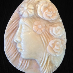 May include: A carved pendant made of pink and white shell, featuring a woman's face in profile with flowers in her hair.