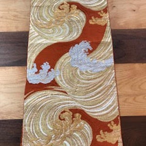 Può includere: Un tessuto rettangolare con un motivo a onde ripetuto in oro, bianco e argento su uno sfondo color ruggine. Il design presenta linee vorticose e creste stilizzate, creando un effetto visivo dinamico. Il tessuto è esposto su una superficie di legno.