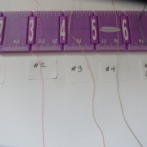 May include: Five different colored threads are laid out on a white surface, labeled #1 Fine to #5 Coarse. The threads are arranged from left to right in order of increasing thickness. A purple ruler is visible at the top of the image.