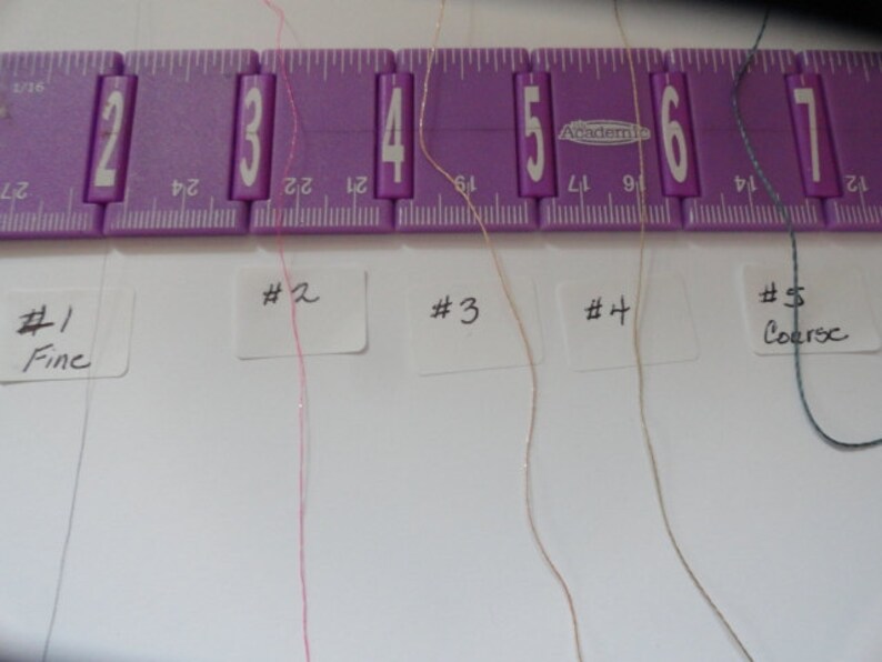 May include: Five different colored threads are laid out on a white surface, labeled #1 Fine to #5 Coarse. The threads are arranged from left to right in order of increasing thickness. A purple ruler is visible at the top of the image.