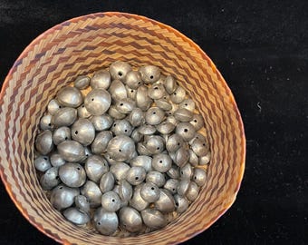 May include: A woven basket filled with silver-colored beads. The beads are various sizes and shapes, with small holes. The basket has a brown and tan woven pattern. The background is a dark, solid color.
