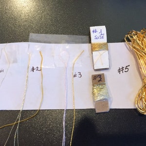 May include: Five spools of gold thread, labelled #1, #2, #3, #3, and #5. The spools are on a white surface with a white background.
